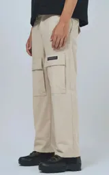 Pantalón cargo de color beige claro, corte recto y holgado, con bolsillos tipo parche en los muslos. Presenta costuras reforzadas en las rodillas y una etiqueta rectangular negra en el bolsillo derecho.