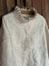 Camisa color natural de lino bordado, de corte oversize, con cuello con volados, cierre frontal con botones y mangas largas.