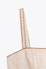 Top corto de escote recto y tirantes finos, con estampado de cuadros vichy en tonos beige y blanco. Presenta tejido elástico tipo nido de abeja en la espalda, detalle de botones forrados en el delantero y bajo con volantes. Cierre con cremallera lateral oculta en la costura.