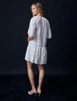 Vestido corto blanco de algodón con bordado calado tipo broderie, cuello redondo, escote en V con lazo y borlas, y mangas tres cuartos con puño.
