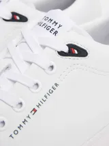 Zapatillas de corte bajo confeccionadas en una mezcla de piel y materiales sintéticos, color blanco. Presentan un diseño atemporal con cordones al tono, suela de goma vulcanizada y detalles distintivos de la marca Tommy Hilfiger, incluyendo el logo en el lateral, la lengüeta y el talón.