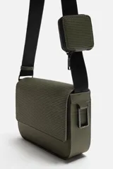 Bandolera negra con acabado engomado y textura acanalada. Tiene un compartimento principal con cierre de solapa y botón a presión, un bolsillo interior con cremallera y un monedero cuadrado en la correa. La correa de hombro es ajustable.