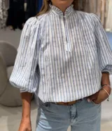 Blusa a rayas verticales celestes y blancas, de manga larga con puños elásticos, cuello alto con volantes y cierre frontal parcial con botones.