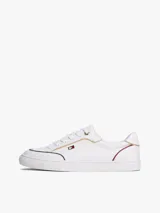 Zapatillas blancas de corte bajo con cordones, estilo urbano y suela cupsole. Presentan ribetes decorativos en azul marino y rojo, detalles metálicos dorados en el cuello y un pequeño logo metálico de Tommy Hilfiger en el lateral.