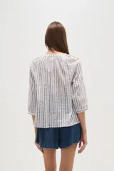 Camisa blanca de algodón con rayas verticales en tono beige/tostado y detalles de bordado tipo ojal a lo largo de las rayas. Presenta cuello mao con abertura en V y botones frontales, y mangas 3/4 con ligero volumen.