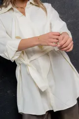 Blusa color crema de poliéster con cuello doble estructurado, volado delantero, lazo regulable en cintura y abotonada al frente.