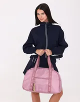 Bolso color beige de tela deportiva con manijas superiores y cierre superior. Tiene un bolsillo frontal con cierre y bolsillos laterales con cierre y tiradores de cordón y goma.