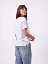 Remera blanca de algodón con cuello redondo y manga corta. Presenta un pequeño bordado en el pecho izquierdo con una figura que parece ser una persona acostada en una tabla de surf o flotador.