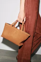 Bolso mini shopper color marrón de serraje, con asa de mano y asa bandolera extraíble. Tiene detalle de pespunte en el corte, bolsillo interior y cierre mediante mosquetón.