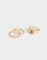 Set de tres anillos dorados de metal. Uno liso, otro con diseño de eslabones entrelazados y otro con forma de sello con una manzana esmaltada y strass.