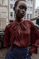 Blusa color marrón con rayas verticales, cuello subido con lazo, escote pico, mangas largas abullonadas con elástico en los puños y cierre frontal con botones forrados.