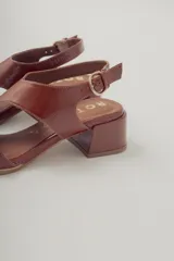 Sandalia de cuero con acabado brillante color marrón rojizo, taco ancho de altura media y punta cuadrada. Presenta tiras anchas cruzadas en el empeine y tira fina con hebilla ajustable en el tobillo.