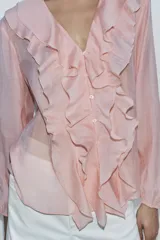 Camisa color rosa pálido confeccionada en tejido ligero con lyocell. Presenta escote en V, cierre frontal con botones, mangas largas abullonadas con puños elásticos y detalle de volantes en el frente.