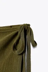 Falda larga color verde oliva, de tiro medio y corte recto. Presenta una superposición de tela tipo pareo ajustable con lazadas.
