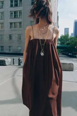 Vestido largo marrón de tejido satinado, con escote recto, tirantes finos extraíbles, bolsillos laterales ocultos y bajo asimétrico acabado en globo.