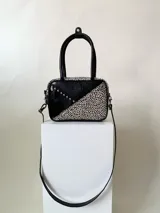 Cartera rígida negra de cuero con tachas plateadas y tres combinaciones de cuero diferentes que aportan textura. Tiene herrajes y cierre metálico plateado marca YKK. Cuenta con un asa fina para llevar al hombro o en la mano y una correa larga extra regulable.