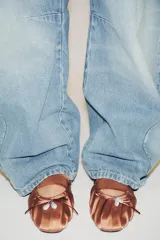 Zapato destalonado plano color marrón, con efecto satinado, detalle de fruncidos en la punta, lazo con perla y tira elástica en el empeine.