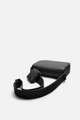 Bandolera negra con acabado engomado y textura acanalada. Tiene un compartimento principal con cierre de solapa y botón a presión, un bolsillo interior con cremallera y un monedero cuadrado en la correa. La correa de hombro es ajustable.