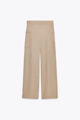 Pantalón de sarga color beige, de tiro medio con cintura elástica en la espalda, bolsillos laterales de plastrón con botón metálico y pierna ancha.