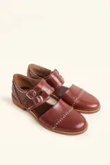 Zapato tipo monk strap con aberturas laterales, confeccionado en cuero vacuno color bordó. Presenta doble tira con hebillas metálicas plateadas en el empeine y suela de cuero con taco bajo.