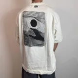 Remera color crudo de corte oversize y mangas cortas, con estampa en la espalda de una mano alcanzando un círculo.