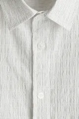Camisa blanca de popelina de algodón texturizado, con cuello inglés, tapeta lisa, canesú con pliegue de caja en la espalda y mangas largas con abertura y cierre ajustable de botón en los puños. Corte holgado y bajo levemente redondeado.