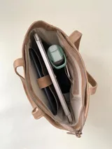 Cartera tipo tote de cuero graneado color negro, con textura uniforme y suave al tacto. Tiene manijas de 55 cm, bolsillo frontal con broche imantado, bolsillo trasero con cierre metálico, cierre principal metálico, forro interior y tres bolsillos internos (uno con cierre, uno plaqué y uno tipo sobre con solapa y botón).