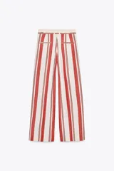 Pantalón de tiro medio con estampado de rayas verticales negras sobre fondo blanco. Tiene trabillas en la cintura, bolsillos laterales, pinzas delanteras y cierre frontal con cremallera, botón y ganchos metálicos.