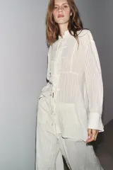 Camisa blanca a rayas verticales, de cuello redondo y manga larga con puños. Presenta cierre frontal con cordones ajustables con lazadas y abertura en la espalda ajustable con lazadas.