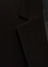 Chaqueta negra con cuello de pico con solapa y cierre de botón en la parte delantera. Tiene manga larga con puños abotonados, diseño recto, estilo traje y dos bolsillos de solapa en la parte delantera.
