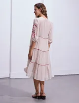Vestido midi blanco con bordados florales azules en el escote, mangas y cintura. Tiene cuello en V, mangas 3/4 con botón y falda escalonada con detalles de ribetes azules.