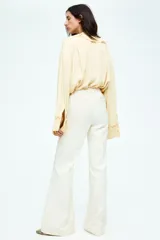 Pantalón acampanado color crema, de tiro bajo, con cierre y pretina prolongada con un botón oculto y uno visible. Modelo con bolsillos laterales discretos, bolsillos atrás insertados y piernas extralargas acampanadas con pliegue cosido adelante y planchada atrás.