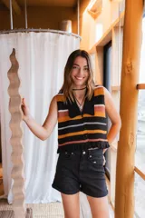 Blusa tejida a crochet con rayas horizontales en colores negro, naranja, blanco y rosa. Tiene cuello camisero, escote en V, botones al frente y ruedo ondulado.