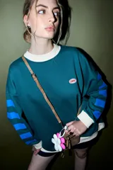 Buzo verde oscuro con cuello redondo y puños y dobladillo en color crema. Presenta un parche ovalado con el texto "CORAL" en el pecho y detalles de franjas azules en las mangas.