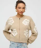 Cardigan cropped color beige con estampado de flores blancas, cuello redondo, cierre frontal con cremallera y mangas largas.