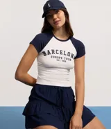 Remera ajustada al cuerpo, estilo raglán, con cuerpo blanco y mangas cortas azul marino. Presenta un estampado en el pecho con el texto "BARCELONA EUROPE TOUR EST. 1992" en azul marino.