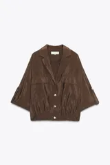 Camisa marrón de cuello solapa y manga corta con trabilla y botón. Tiene bolsillos delanteros de plastrón con solapa, bajo elástico y cierre frontal con botones a presión.