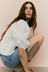 Blusa blanca de algodón con textura de lunares. Tiene escote redondo con lazo, botones en el frente, mangas tres cuartos abullonadas, cordón ajustable en la cintura y peplum.