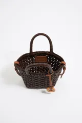 Bolso tipo tote de mimbre color marrón oscuro, con doble asa corta y correa de cuero marrón. Incluye dije circular dorado con letras grabadas.