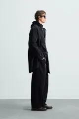 Parka negra de tejido técnico ligero, corte regular, cuello alto con capucha ajustable, manga larga con puños ajustables, cintura ajustable con cordón interno, bolsillos de solapa en la cadera, abertura en la espalda y cierre frontal con cremallera oculta y botones a presión.