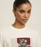Remera blanca de algodón con cuello redondo y mangas cortas. Presenta un estampado rectangular con un diseño de look fashion que incluye un pantalón animal print de vaca, una cartera roja y el texto "Greetings From New York City".