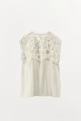 Blusa sin mangas color blanco con cuello alto, bordados y puntillas combinadas a tono, y apliques de abalorios. Cierre frontal con abertura, lazada y botón.