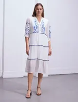 Vestido midi blanco con bordados florales azules en el escote, mangas y cintura. Tiene cuello en V, mangas 3/4 con botón y falda escalonada con detalles de ribetes azules.