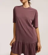 Vestido tipo camiseta de algodón, color malva o ciruela con efecto lavado, de corte recto y holgado, con cuello redondo y mangas cortas. Presenta un volado fruncido en el bajo.