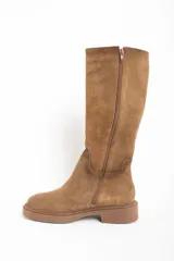 Bota de caña alta color camel, confeccionada en gamuza. Presenta una suela de goma gruesa y plana, y un diseño liso sin cierres visibles.
