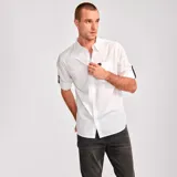 Camisa de popelina verde con cuello abotonado, cierre frontal con botones, bolsillo de parche en el pecho izquierdo, mangas largas con trabillas para enrollar y dobladillo camisero.