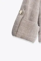 Chaqueta jaspeada color beige, con cuello solapa, manga tres cuartos con trabilla y botón, bolsillos delanteros de plastrón con solapa, bajo ajustable con cordones interiores y cierre frontal con botones a presión.