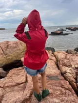 Piloto impermeable de color rojo con detalles de volados verdes en los hombros. Presenta capucha integrada, cintura ajustable con cordón, mangas con volumen y puños elastizados.