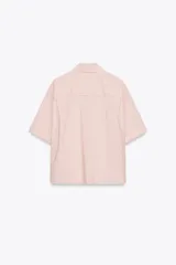 Camisa color rosa claro a rayas verticales finas color marrón, de popelín, con cuello solapa y manga corta con dobladillo. Tiene un bolsillo de parche en el pecho, aberturas laterales en el bajo y pinzas en la espalda. El cierre frontal es con botones ocultos.
