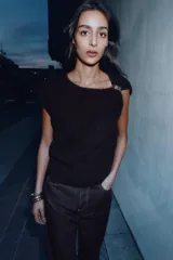 Jersey de punto negro, sin mangas (manga sisa), con cuello cuadrado drapeado y un broche dorado decorativo en el hombro.